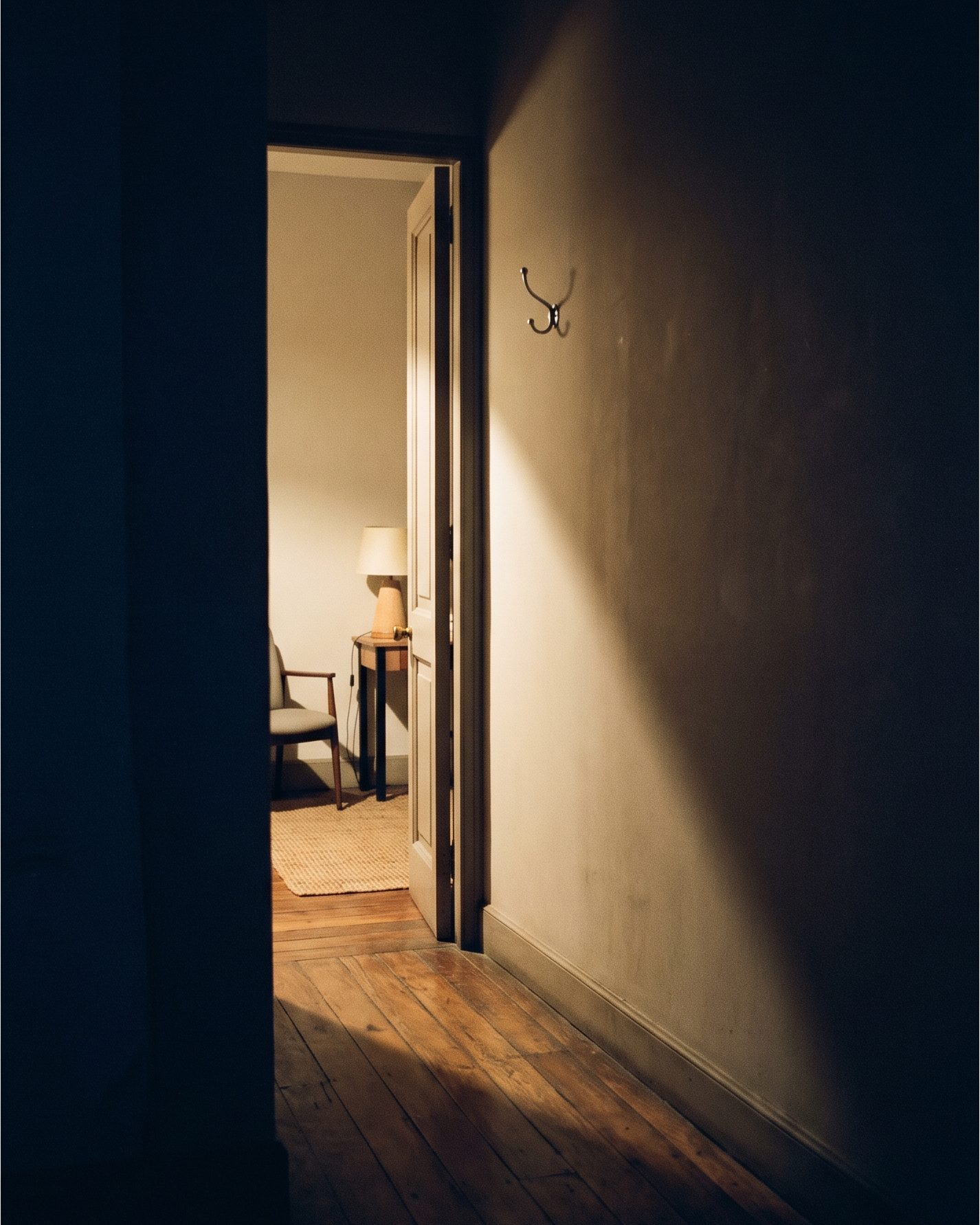 Puerta abierta vista desde un pasillo a oscuras en hora azul, con la luz cálida del cuarto de al lado cayendo en cuña sobre el piso de madera.