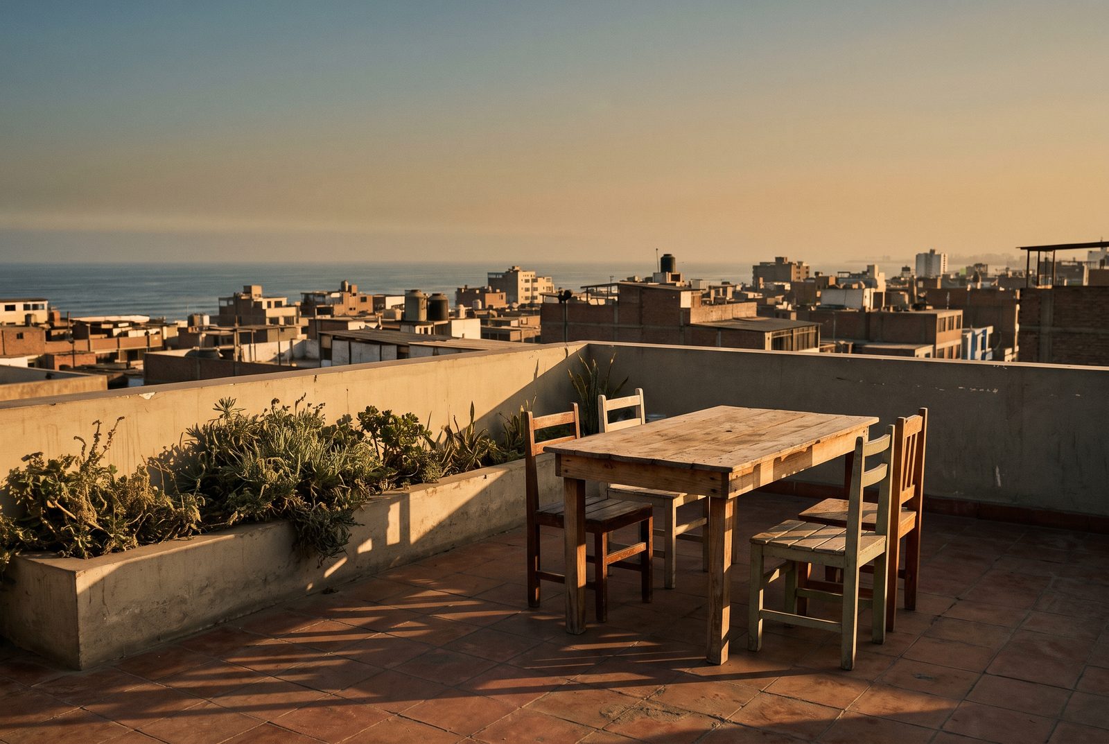 Azotea sobre un distrito de Lima en hora dorada, con una mesa de madera y cuatro sillas sobre losetas de terracota y, al fondo, los techos de la ciudad hasta el mar.